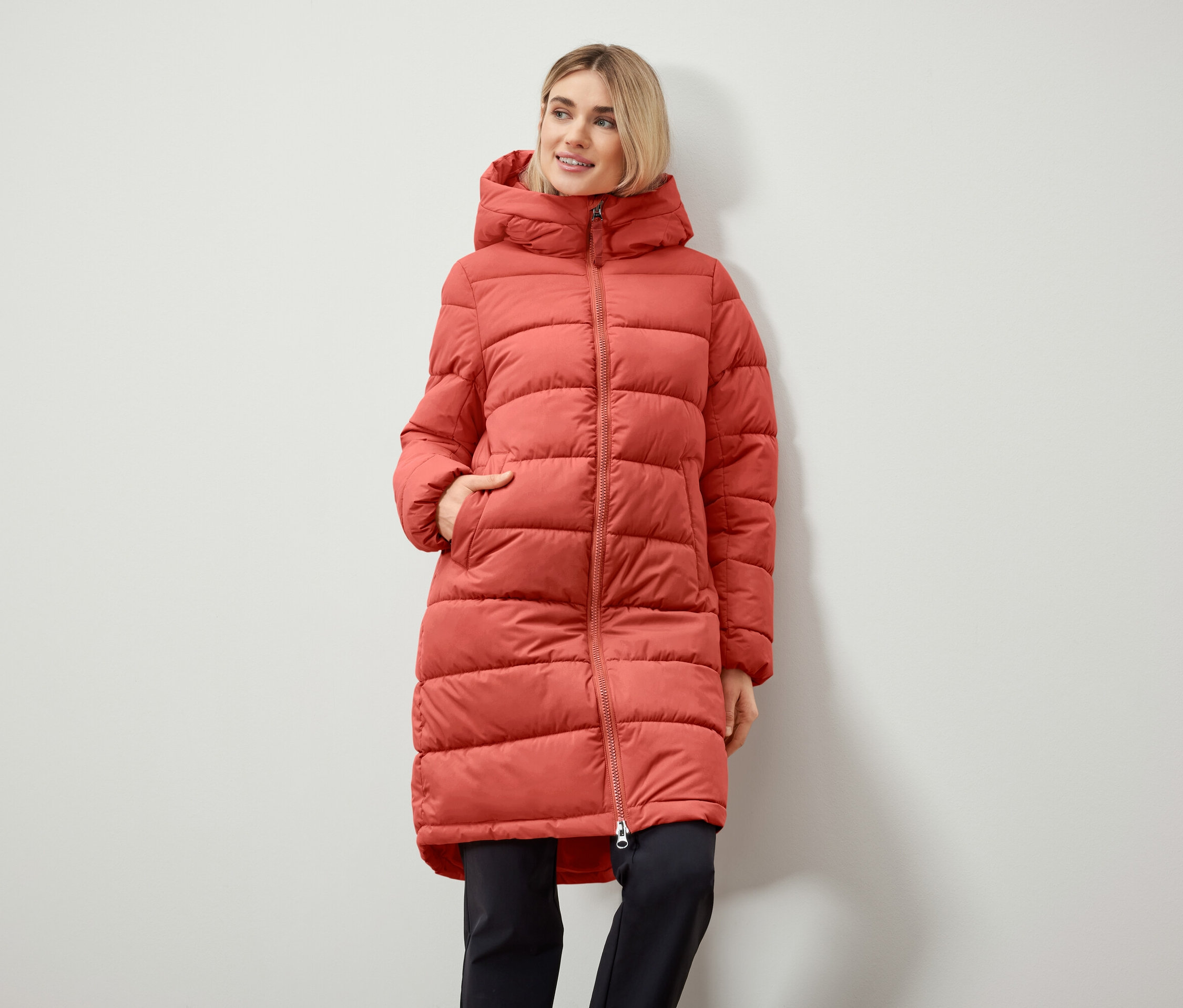 Kadın kapüşonlu uzun şişme mont giyiyor ve elleri cebinde.