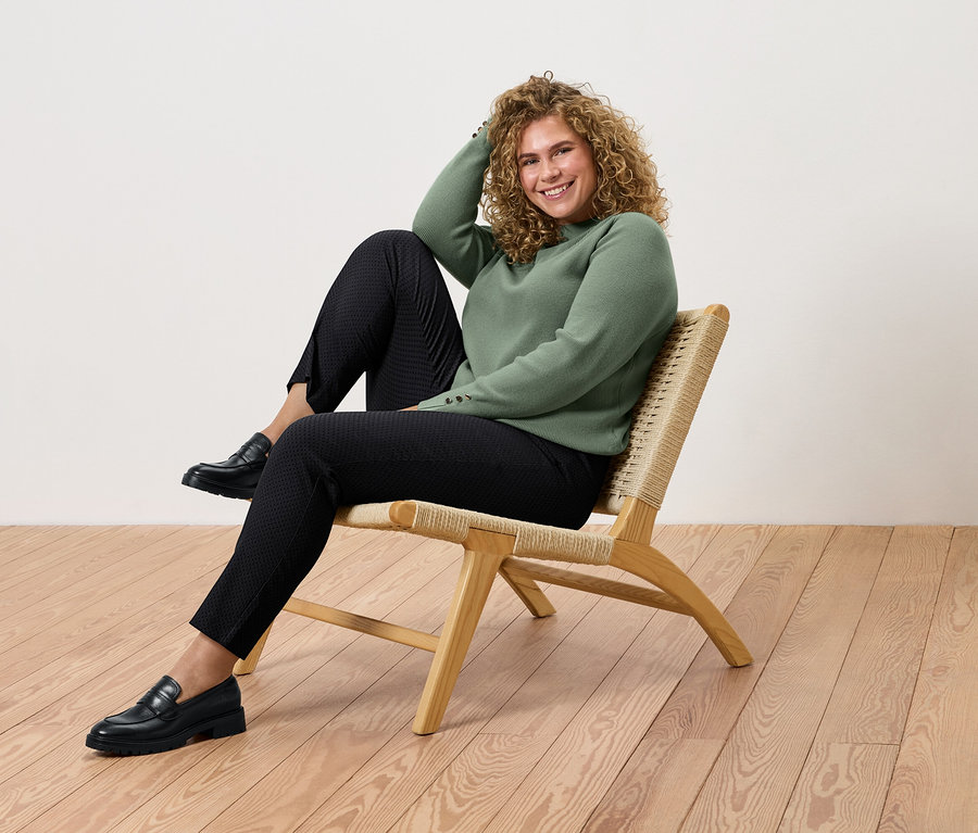 Kıvırcık saçlı bir kadın rattan bir sandalyede oturuyor, yeşil bir kazak, siyah pantolon ve ayakkabılar giyiyor.