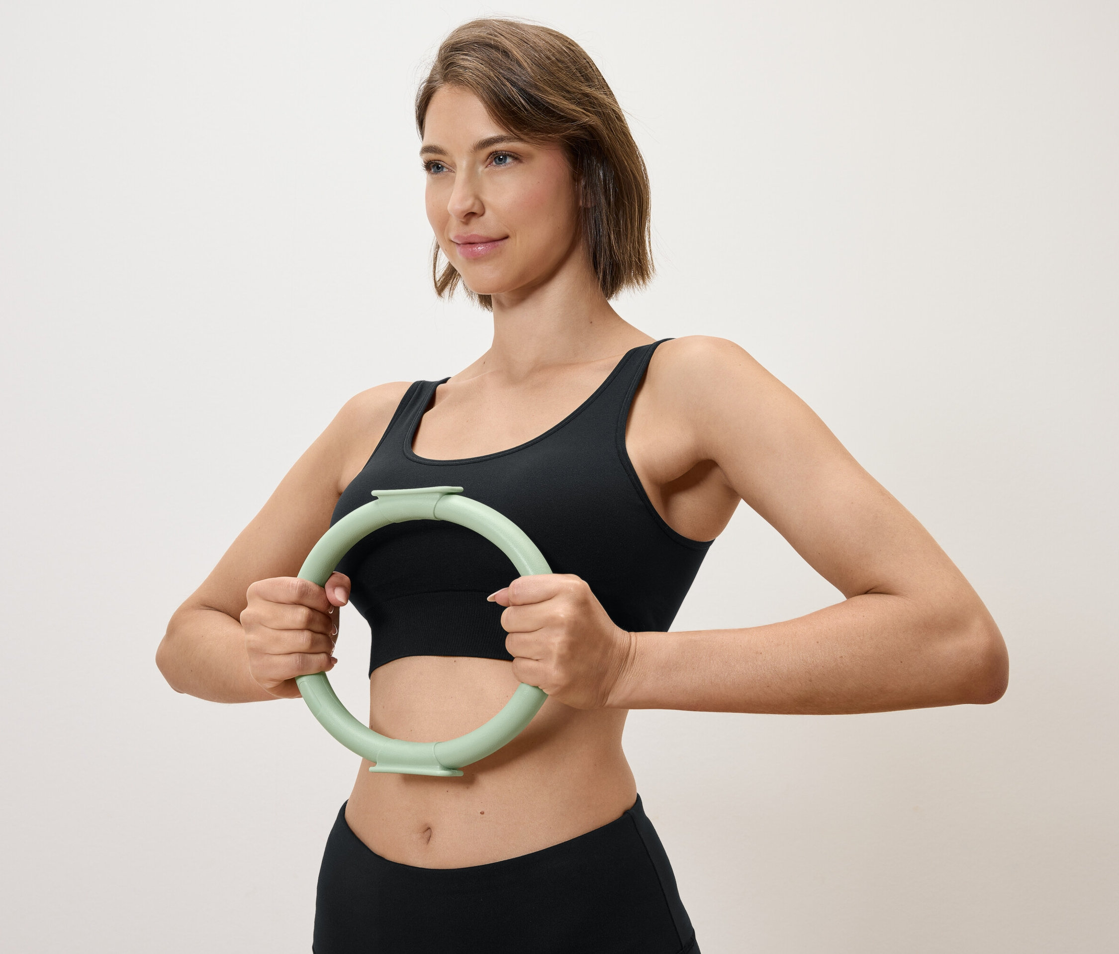 Kadın, siyah spor üstü ve tayt giyerken açık yeşil bir egzersiz çemberini önünde tutuyor.
