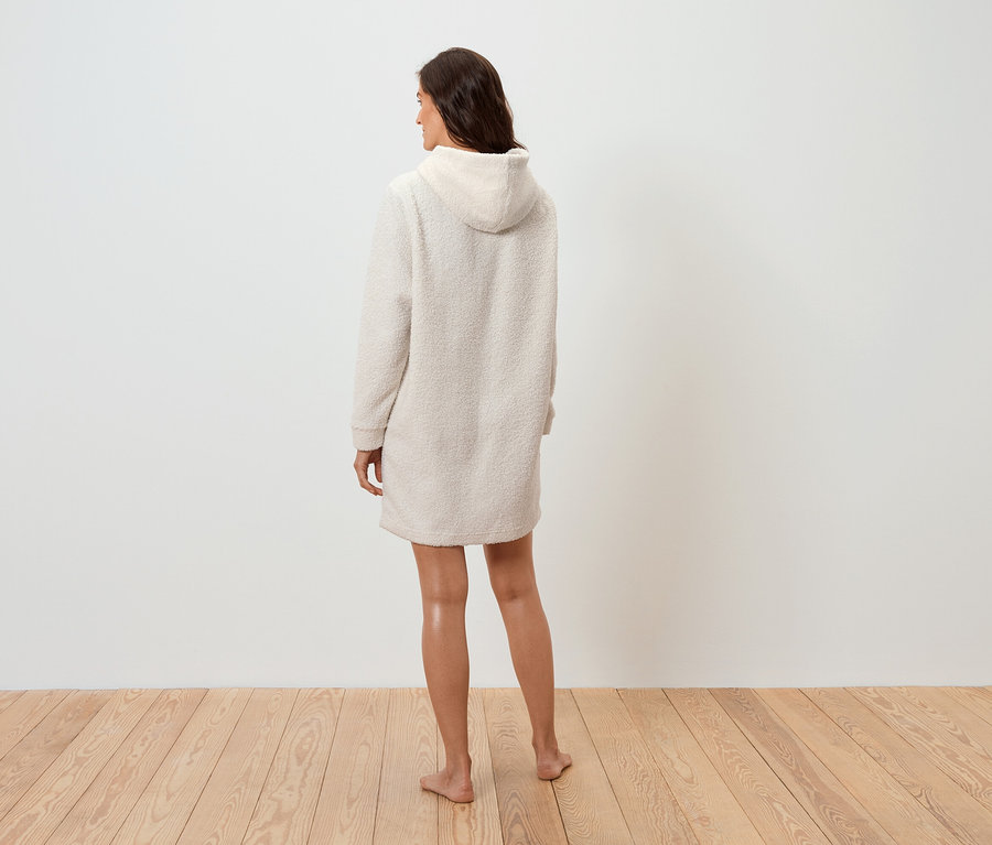 Krem rengi kapüşonlu bir elbise giyen bir kadın, ahşap bir zeminde kameraya arkası dönük duruyor.