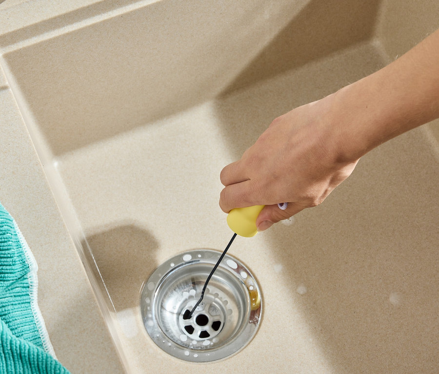 Bir el, sarı bir aletle lavabo giderini temizliyor. Lavabonun yanında yeşil bir bez var.