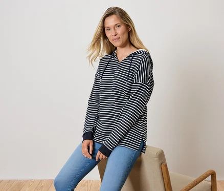 Bir kadın sandalyede oturuyor, mavi kot pantolon ve çizgili kapüşonlu bir sweatshirt giyiyor.