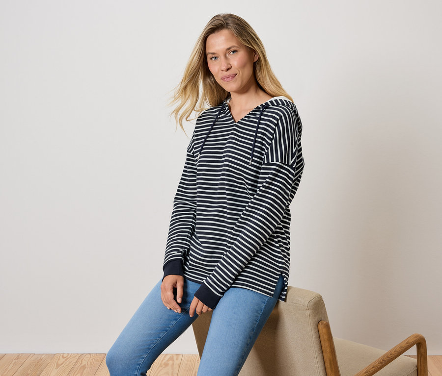 Bir kadın sandalyede oturuyor, mavi kot pantolon ve çizgili kapüşonlu bir sweatshirt giyiyor.