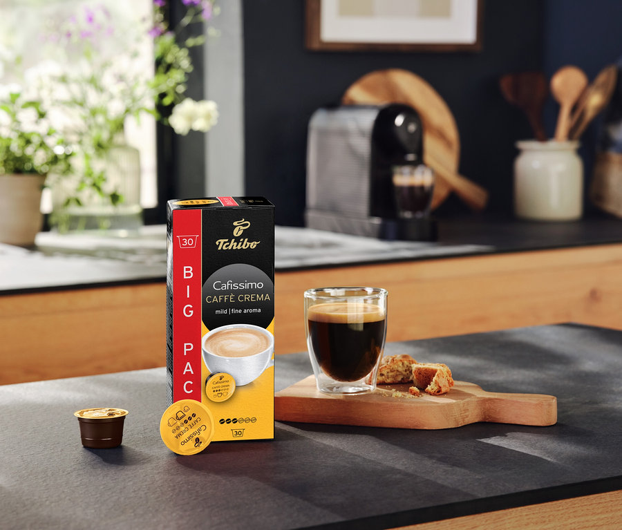 Mutfak tezgahında bir paket Cafissimo Caffè Crema mild kapsül, bir fincan kahve ve bir kahve makinesi.