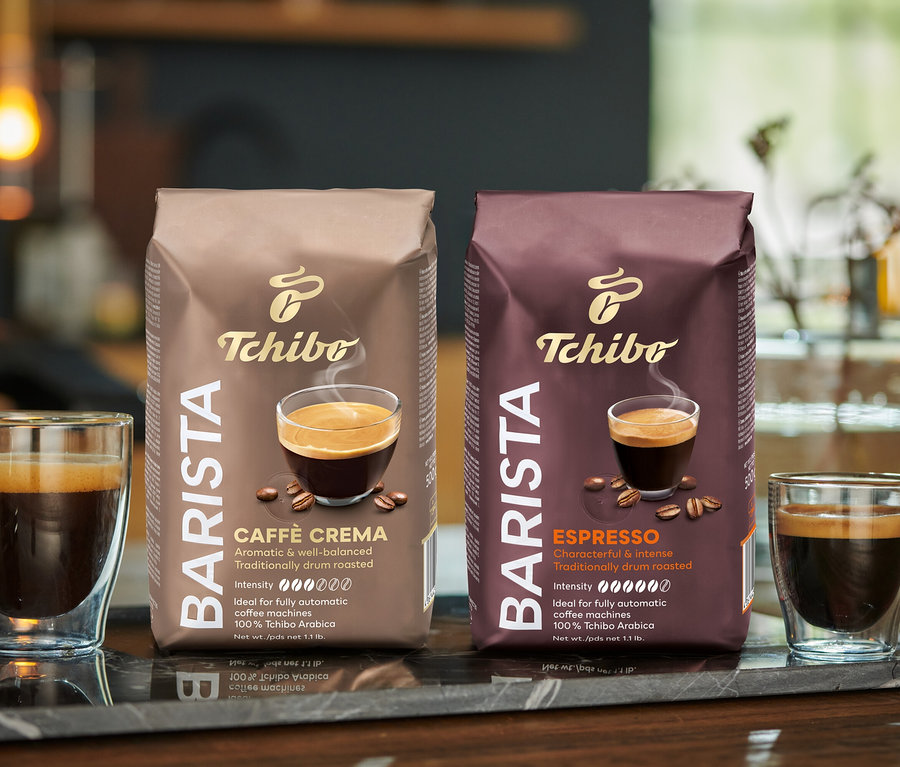 İki bardak kahve ve iki paket Tchibo BARISTA Caffè Crema Çekirdek Kahve, mermer bir yüzey üzerinde duruyor.