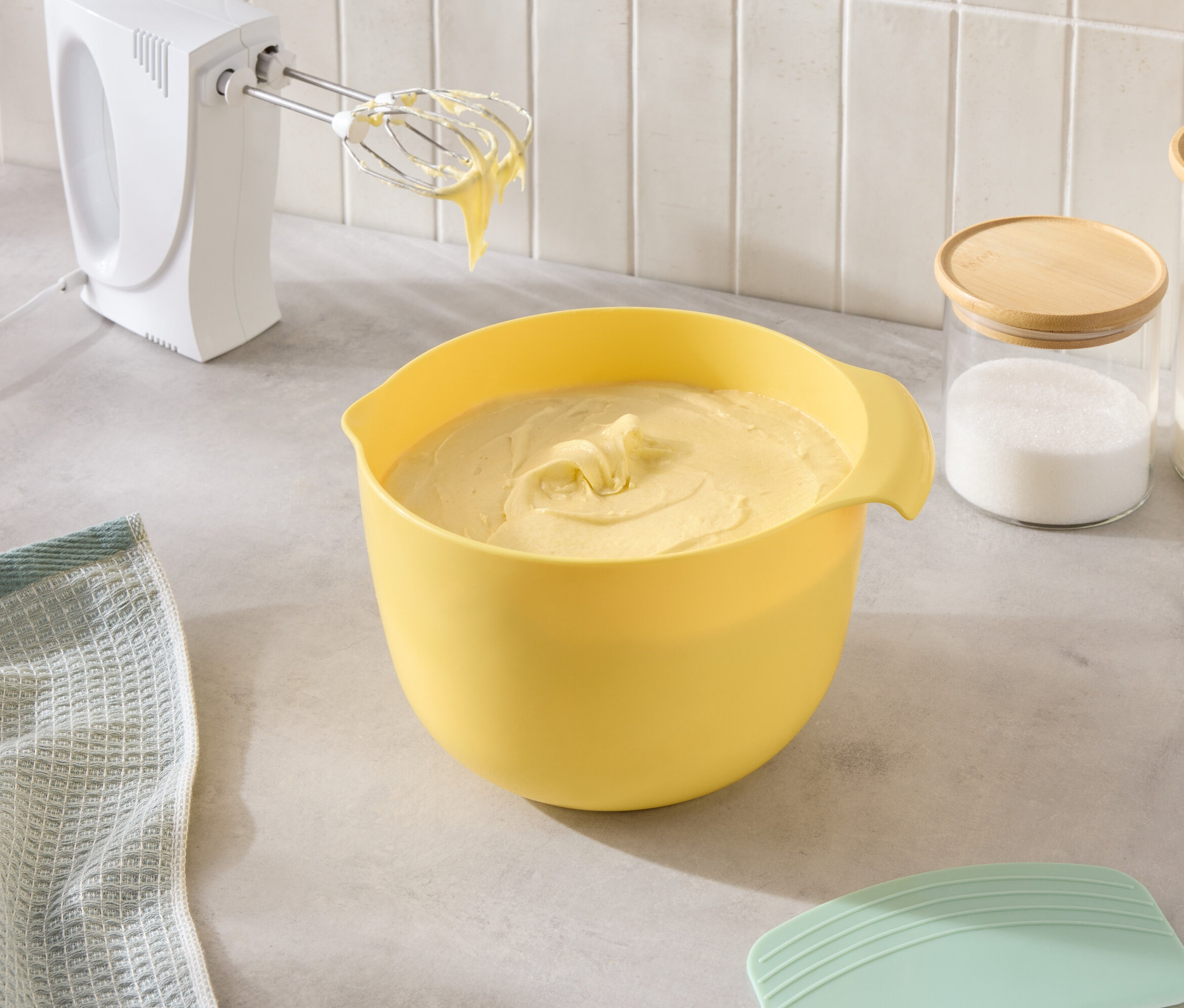 Tezgahta sarı bir hamur kabı, bir mikser ve şeker ve un kavanozları duruyor.
