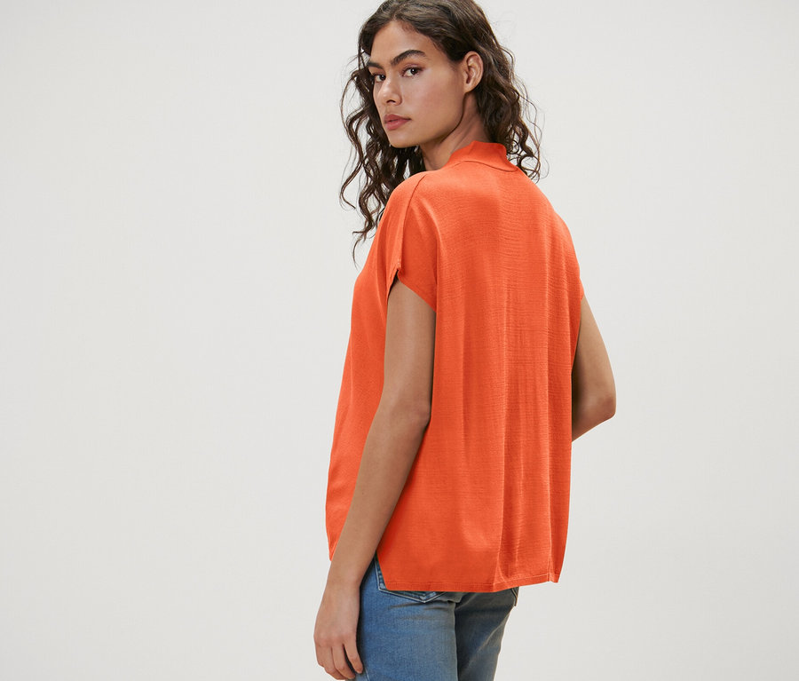 Kadın omzunun üzerinden bakıyor, turuncu, kısa kollu bir hırka ve kot şort giyiyor.