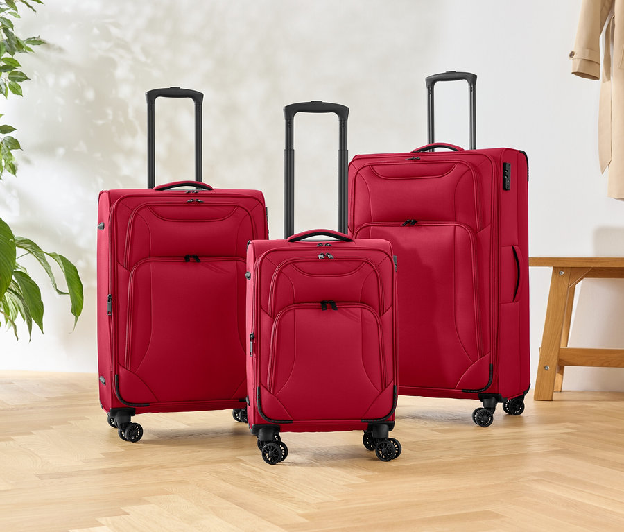 Farklı boyutlarda üç adet tekerlekli kırmızı bavul, ahşap bir zemin üzerinde duruyor, yanında bir bitki ve bir bank var.