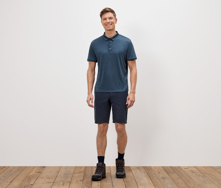 Bir adam beyaz bir duvarın önünde duruyor, mavi polo yaka tişört, kısa şort ve yürüyüş ayakkabıları giyiyor.