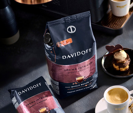 Bir masa üzerinde iki paket Davidoff Crema Intense kahve çekirdeği, bir fincan kahve ve bir tatlı.