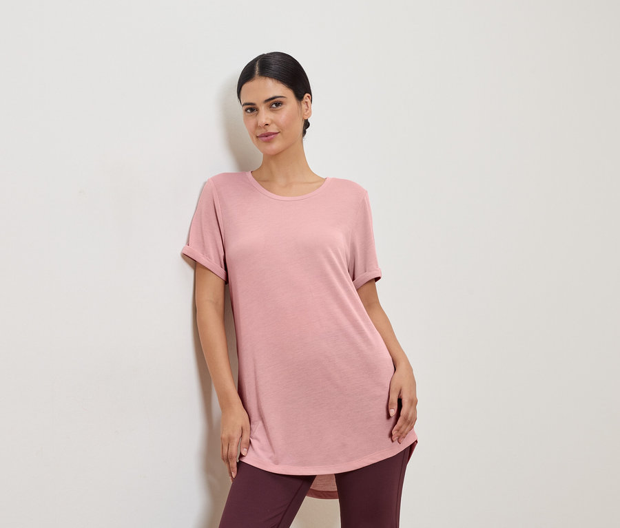 Pembe tişört ve tayt giyen bir kadın beyaz bir duvara yaslanıyor.