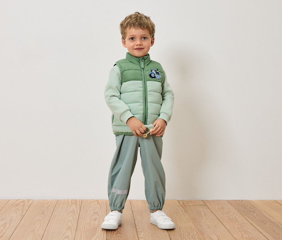 Açık yeşil ceket, açık yeşil pantolon ve beyaz spor ayakkabı giyen bir çocuk duvarın önünde duruyor.