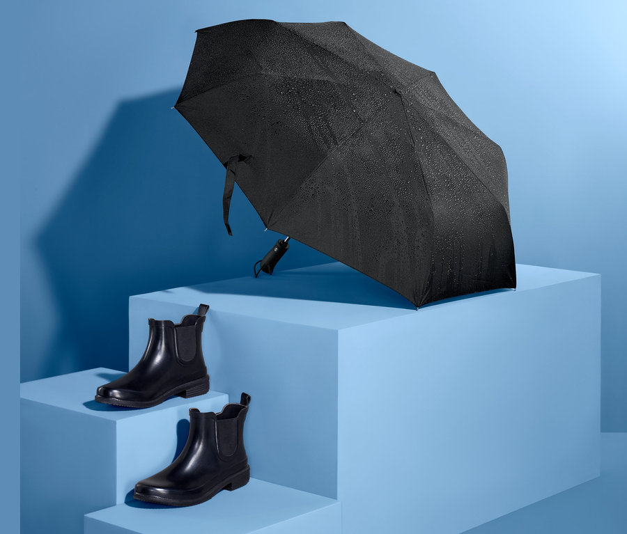 Mavi bir kaide üzerinde siyah otomatik cep şemsiyesi ve yanında bir çift siyah ayakkabı.