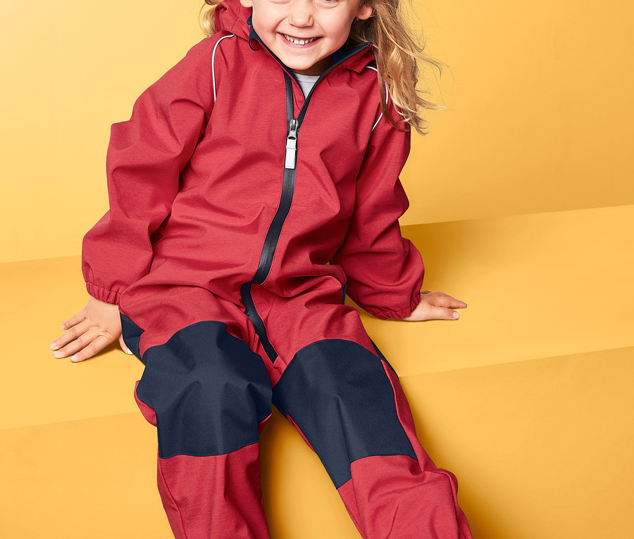 Gülümseyen bir kız sarı bir arka planda oturuyor ve softshell tulum giyiyor.