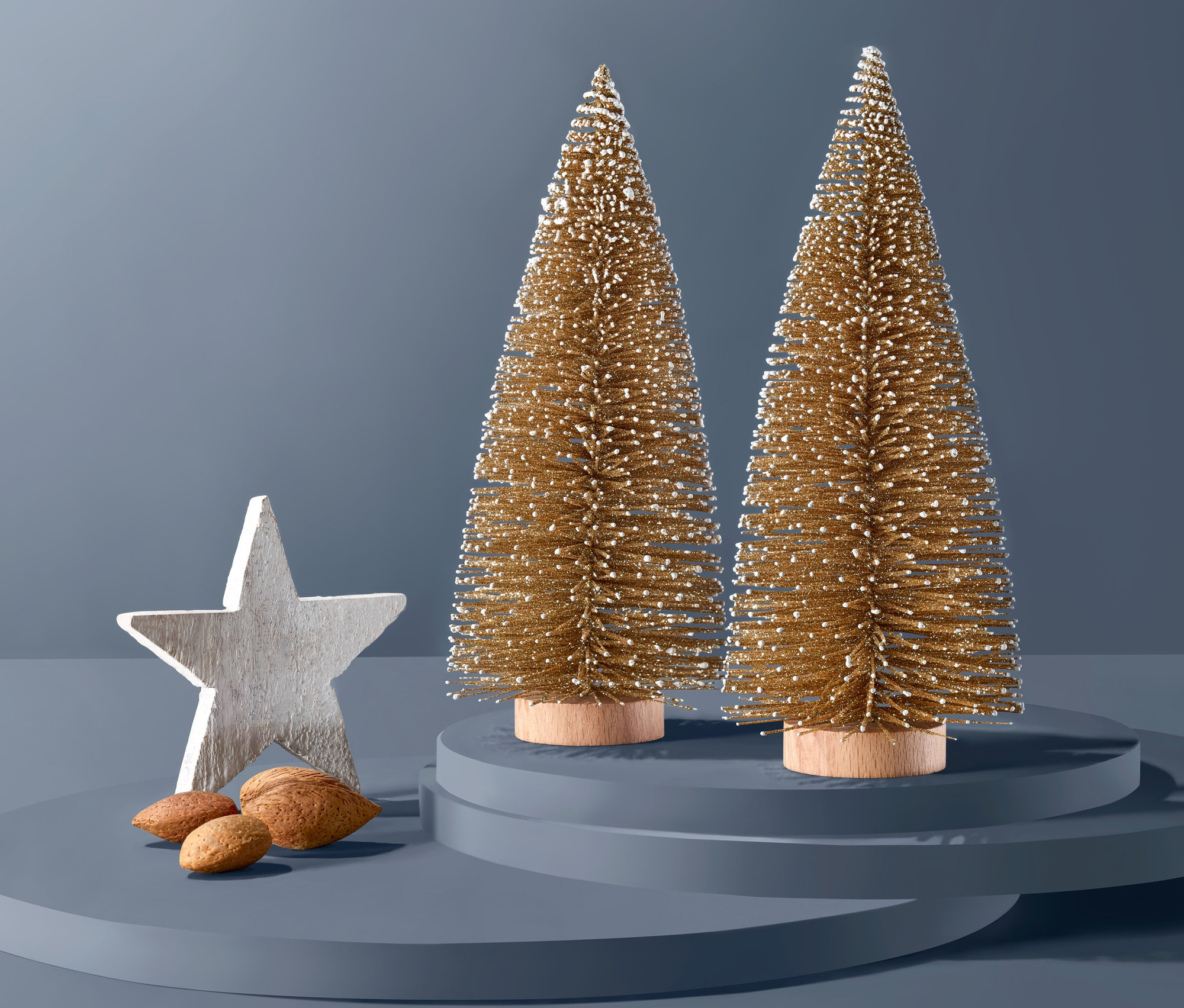 İki altın renkli dekoratif ağaç, beyaz bir yıldız ve bademlerin yanında mavi bir kaide üzerinde duruyor.