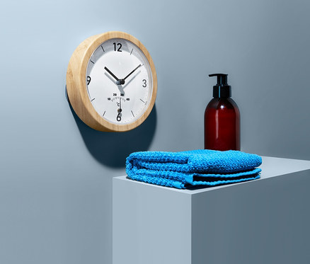 Duvara termometreli banyo duvar saati asılı. Bir kaide üzerinde mavi bir havlu ve pompalı kahverengi bir şişe duruyor.