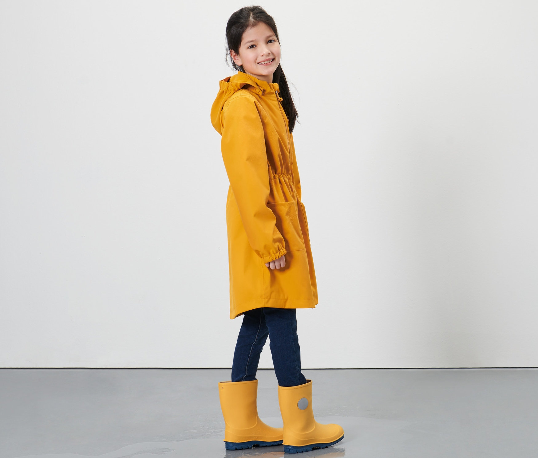 Gülümseyen kız çocuk sarı yağmur parkası ve sarı lastik çizme giyiyor, yandan görünüm.