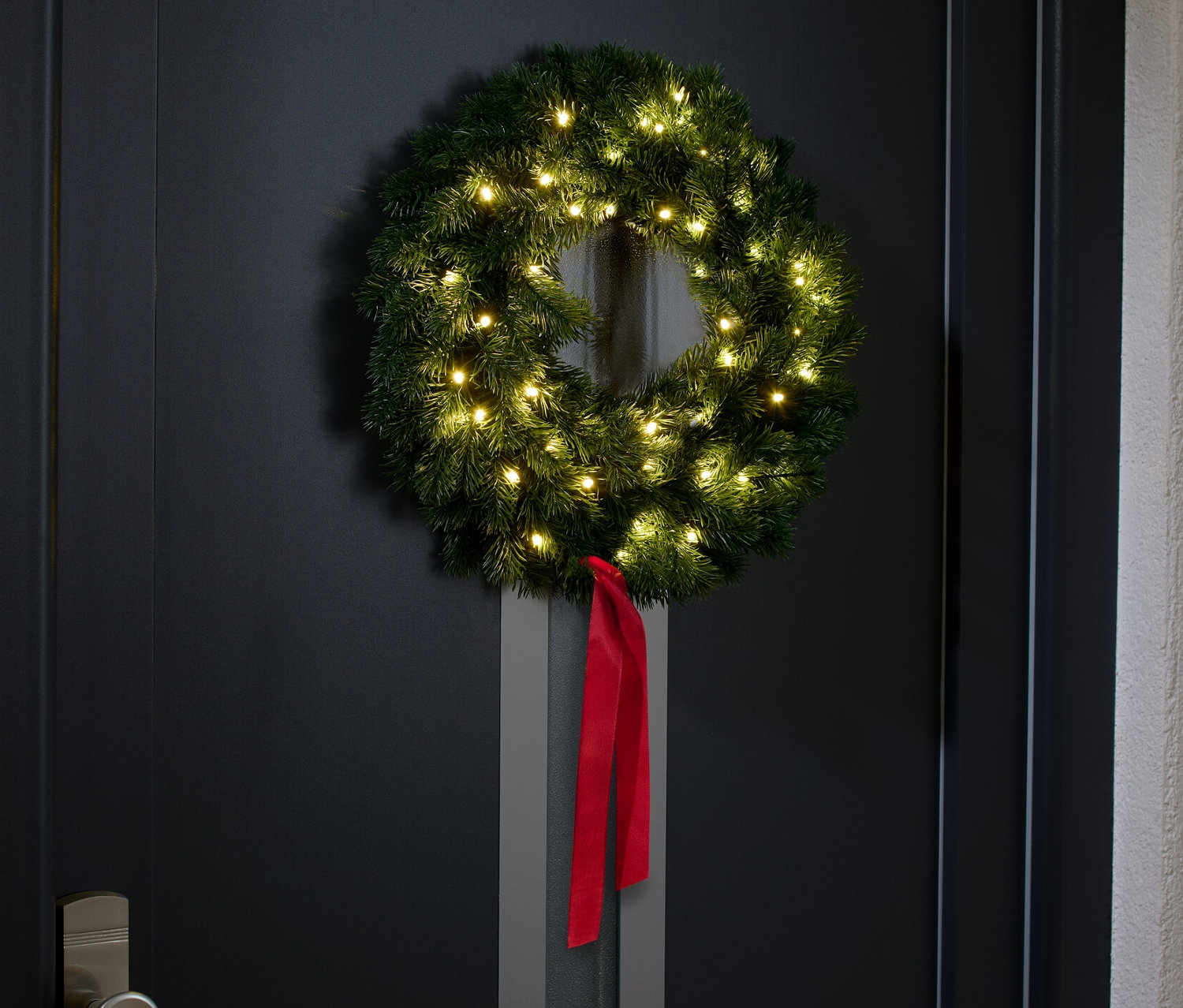 Siyah bir kapıda kırmızı kurdele ile süslenmiş LED'li Çam Girlant asılı duruyor.