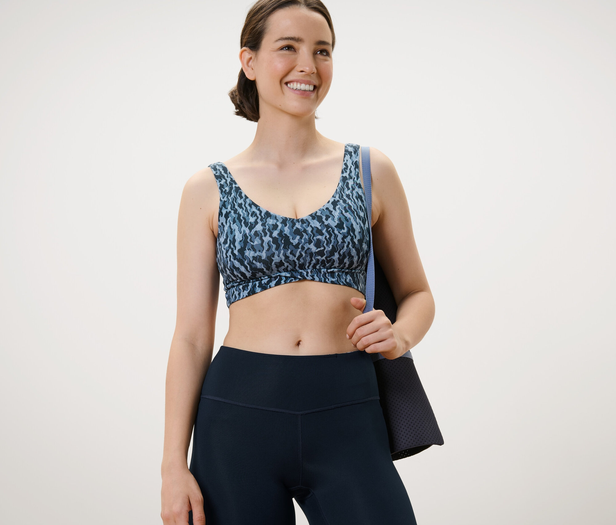 Kadın, tamamı baskılı spor büstiyer giyiyor ve bir egzersiz matı tutuyor.