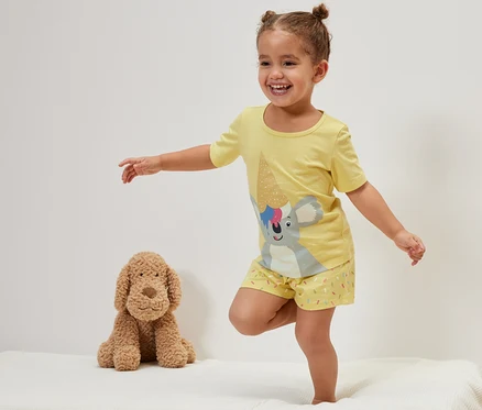 Gülümseyen küçük bir kız, peluş bir köpeğin yanında yatakta duruyor ve sarı Çocuk Şortlu Pijama Takımı, Koala giyiyor.