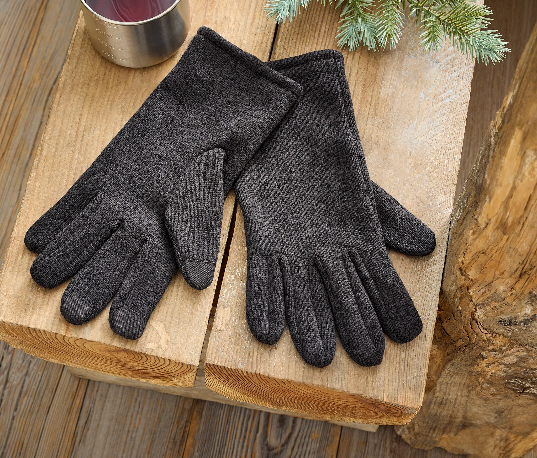 Bir çift gri eldiven, gümüş rengi bir bardak ve bir çam dalının yanında ahşap bir yüzeyde duruyor.