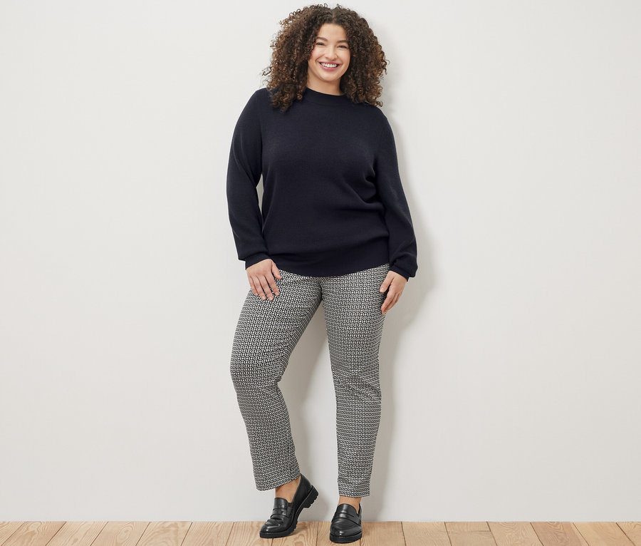 Gülümseyen bir kadın lacivert ince örgü kazak ve baskılı streç pantolonla poz veriyor.