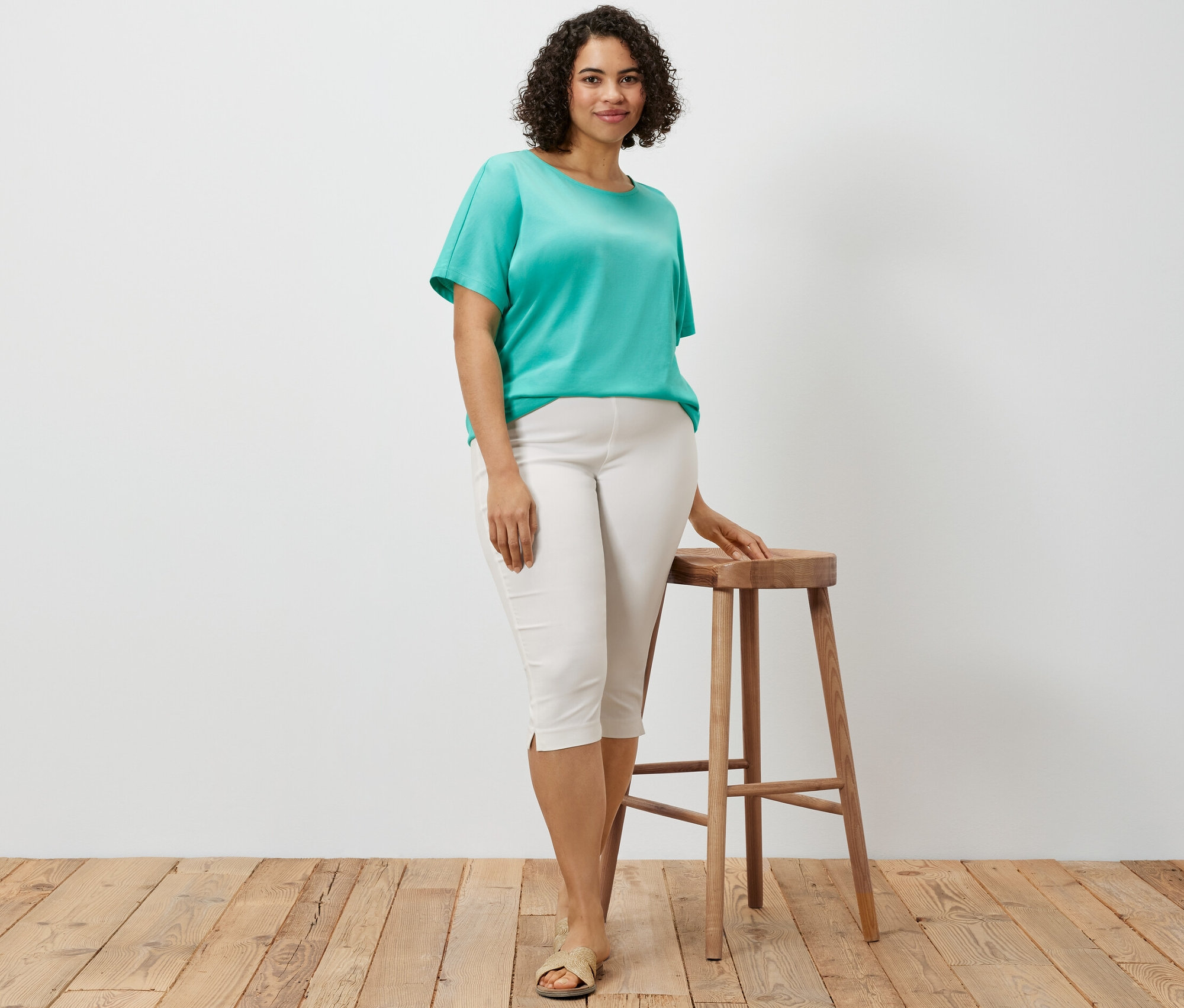 Kadın ahşap bir tabureye yaslanmış, beyaz 3/4 streç pantolon ve deniz mavisi düşük omuzlu tişört giyiyor.