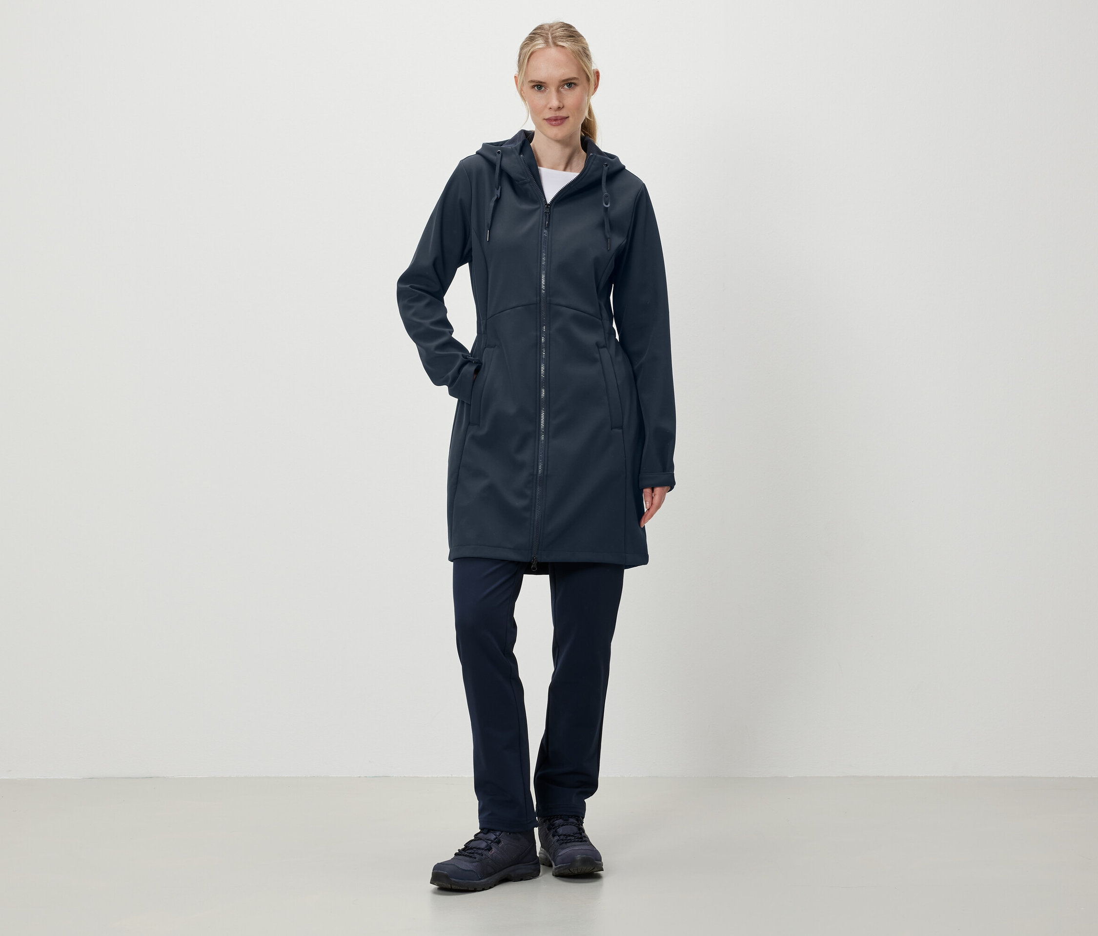Bir kadın ayakta duruyor ve lacivert kapüşonlu bir mont, pantolon ve ayakkabı giyiyor.