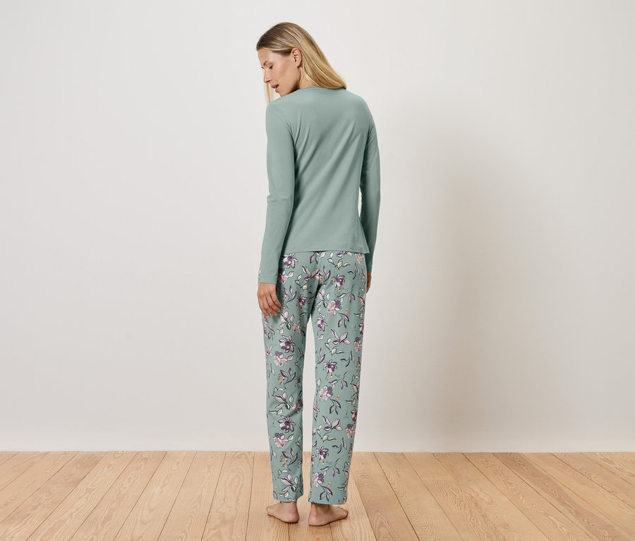 Kadın kameraya arkası dönük duruyor, uzun kollu açık yeşil bir pijama ve çiçekli bir pantolon giyiyor.