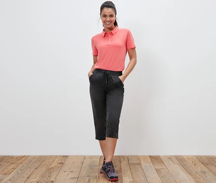 Bir kadın mercan rengi polo yaka tişört ve fonksiyonel softshell kapri ile ayakta poz veriyor.