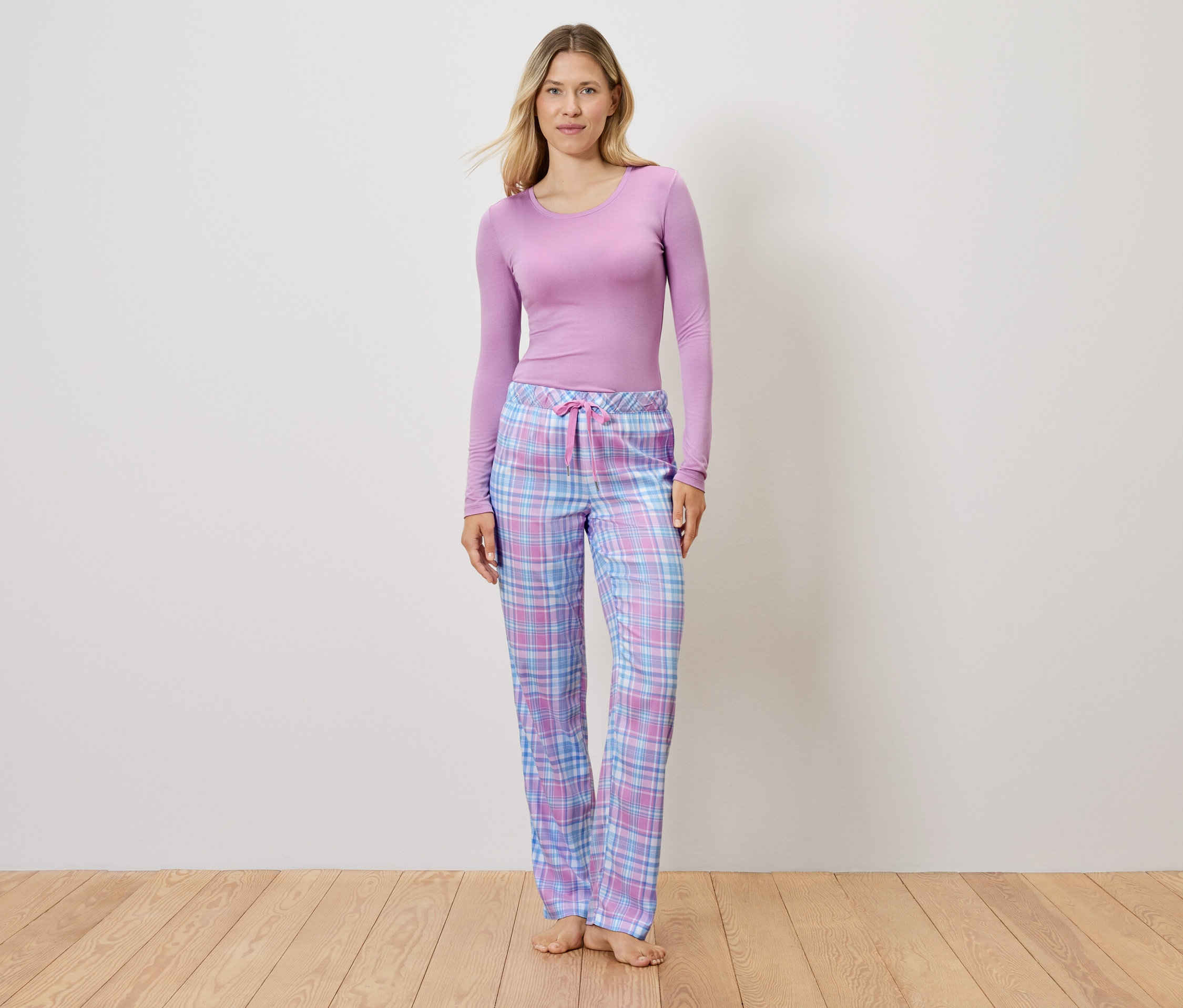 Lila bir üst ve ekose pijama pantolonu giyen bir kadın ahşap bir zeminde duruyor.