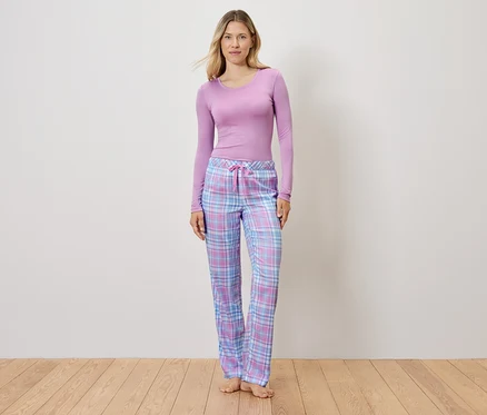 Lila bir üst ve ekose pijama pantolonu giyen bir kadın ahşap bir zeminde duruyor.