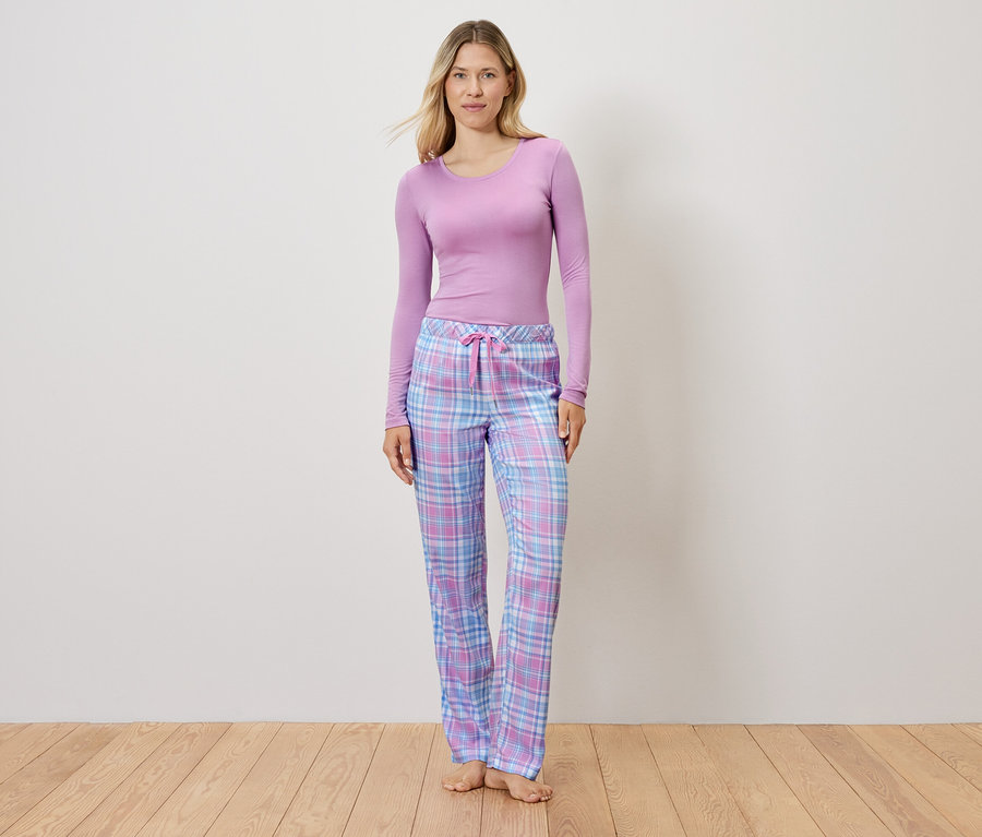 Lila bir üst ve ekose pijama pantolonu giyen bir kadın ahşap bir zeminde duruyor.