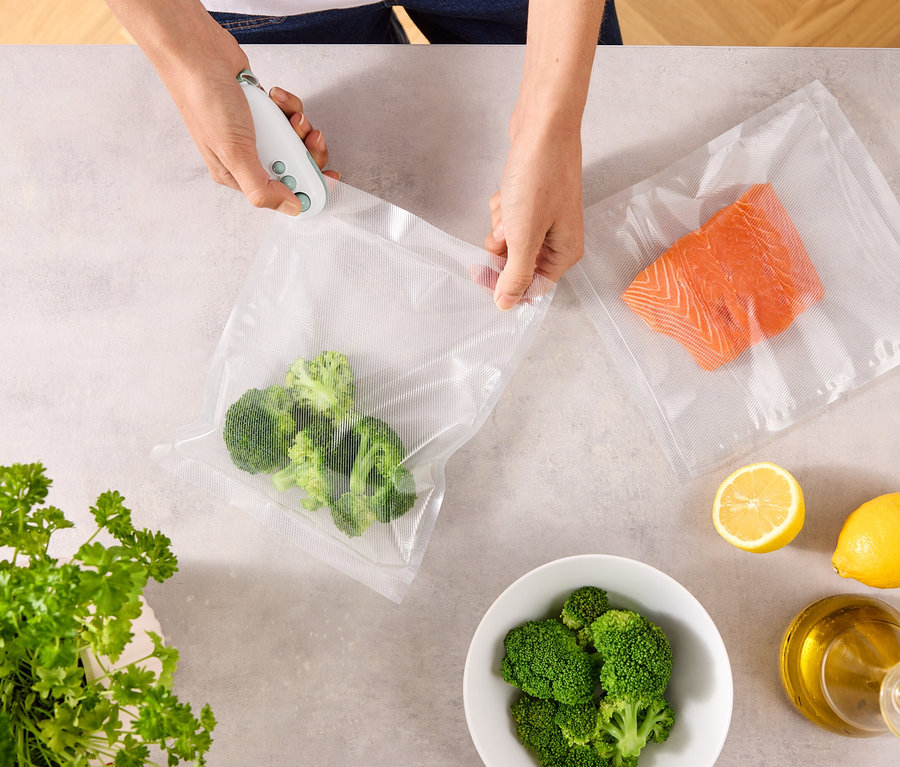 Bir kişi vakum makinesiyle brokoli dolu bir poşeti kapatıyor. Masada somonlu başka bir poşet, limonlar, bir kase brokoli, maydanoz ve bir şişe yağ var.