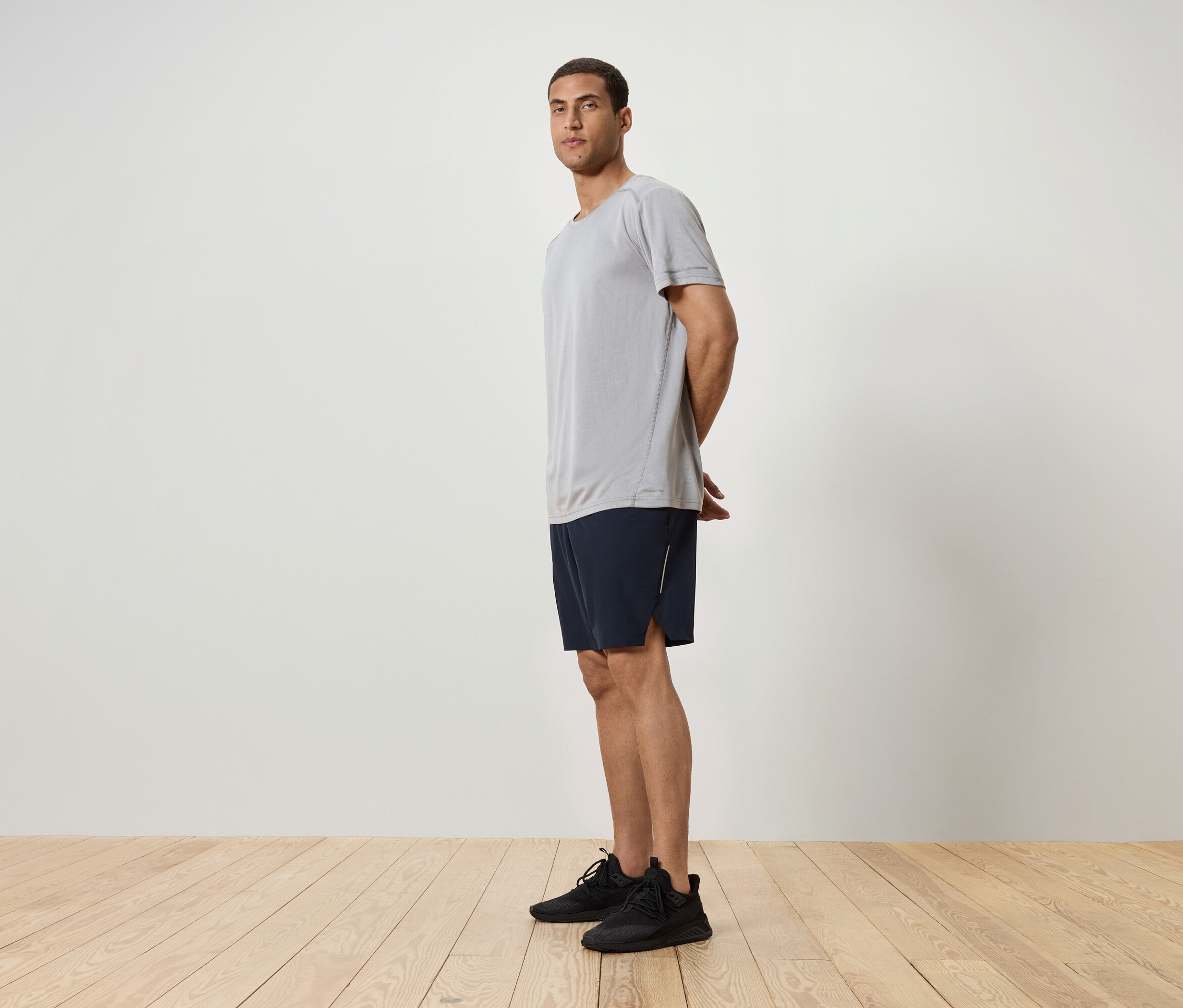 Adam yandan duruyor, gri bir tişört, lacivert şort ve siyah spor ayakkabı giyiyor.