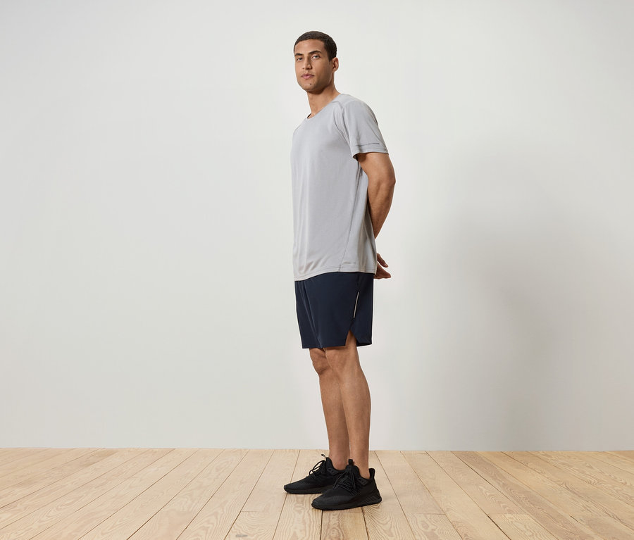 Adam yandan duruyor, gri bir tişört, lacivert şort ve siyah spor ayakkabı giyiyor.