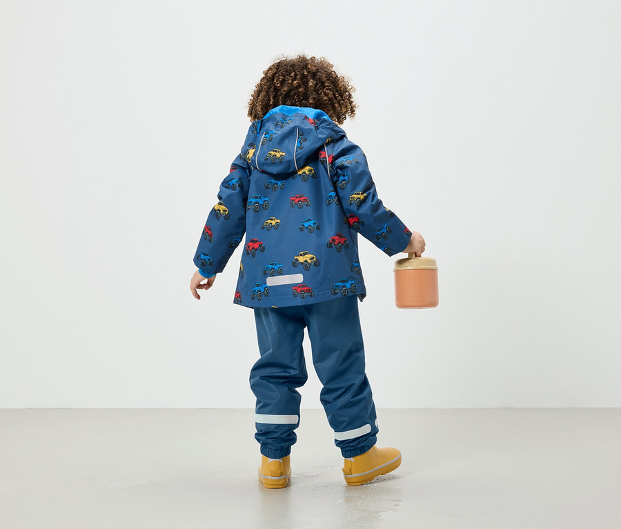 Araba baskılı mavi ceketli çocuk, turuncu bir kap tutuyor.