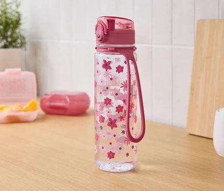 Ahşap bir masanın üzerinde pembe çiçekli, pembe kapaklı ve askılıklı şeffaf bir su şişesi duruyor. Arka planda mandalinalı bir yemek kutusu, kapalı bir kutu, bir kesme tahtası ve bir şekerlik var.