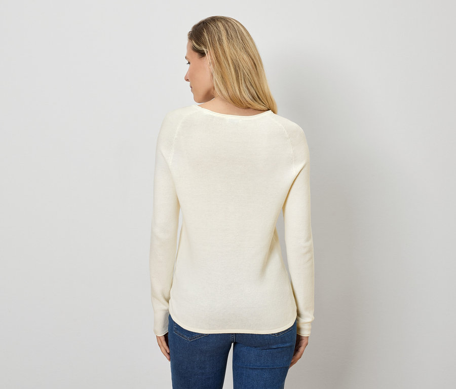 Sarışın saçlı bir kadın kameraya arkası dönük duruyor, üzerinde krem rengi bir kazak ve kot pantolon var.