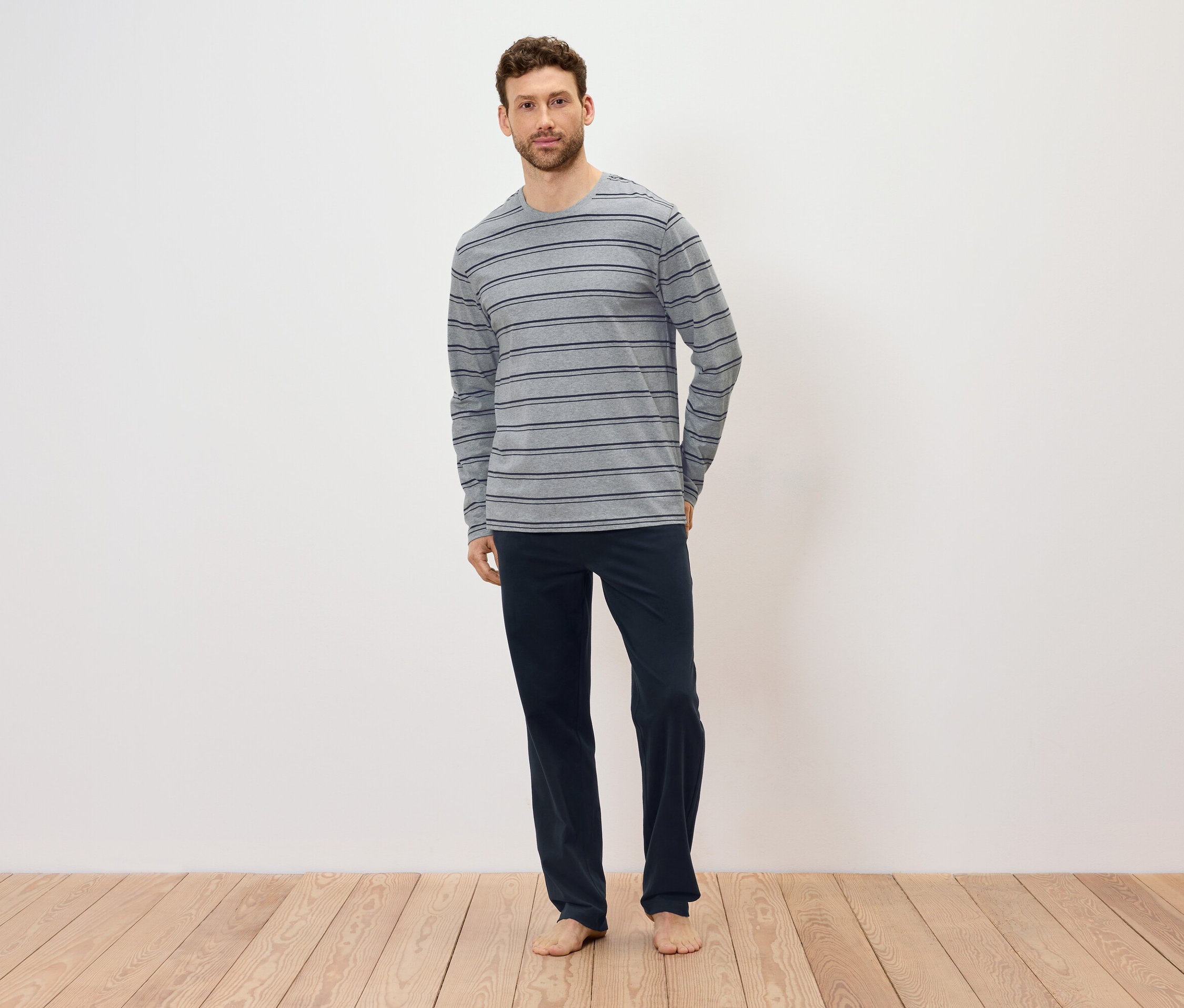 Adam ahşap zeminde ayakta duruyor, üzerinde gri çizgili uzun kollu bir tişört ve lacivert pantolon var.