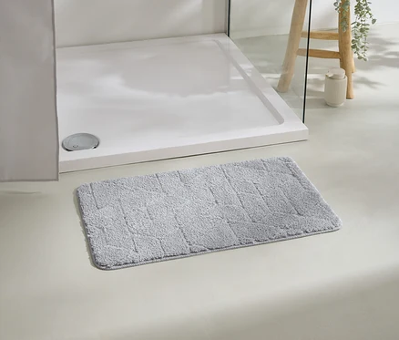 Beyaz duş teknesinin önünde gri bir banyo paspası duruyor, arka planda bitki olan bir tabure.