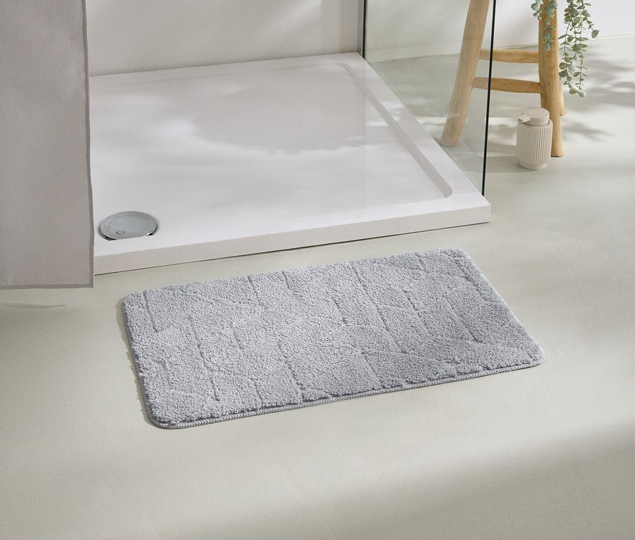 Beyaz duş teknesinin önünde gri bir banyo paspası duruyor, arka planda bitki olan bir tabure.
