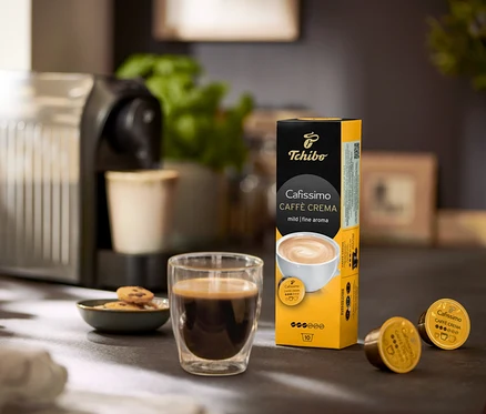 Bir masanın üzerinde kahve makinesi, bir bardak kahve ve bir paket Caffè Crema Fine Aroma– 120 Kapsül bulunmaktadır.