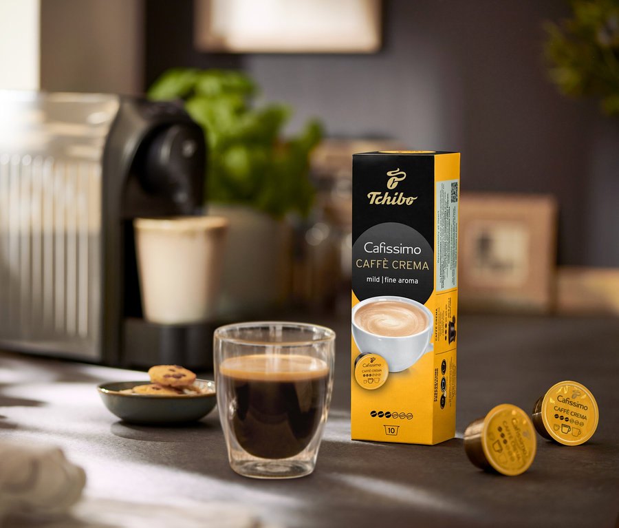 Bir masanın üzerinde kahve makinesi, bir bardak kahve ve bir paket Caffè Crema Fine Aroma– 120 Kapsül bulunmaktadır.