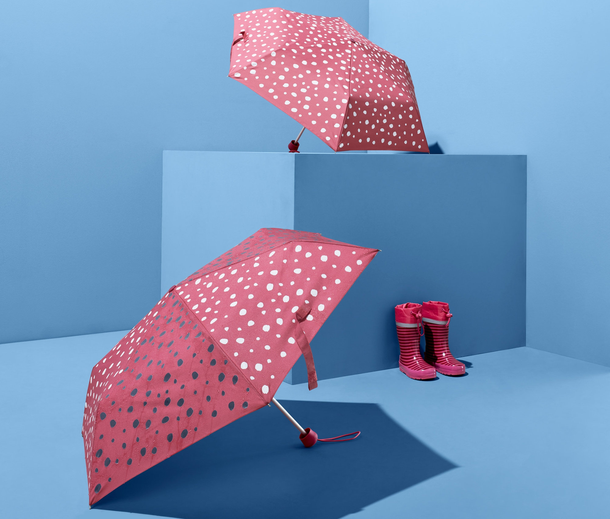 Mavi bir arka planda iki adet Renk Değiştiren Çocuk Cep Şemsiyesi ve bir çift çocuk çizmesi.