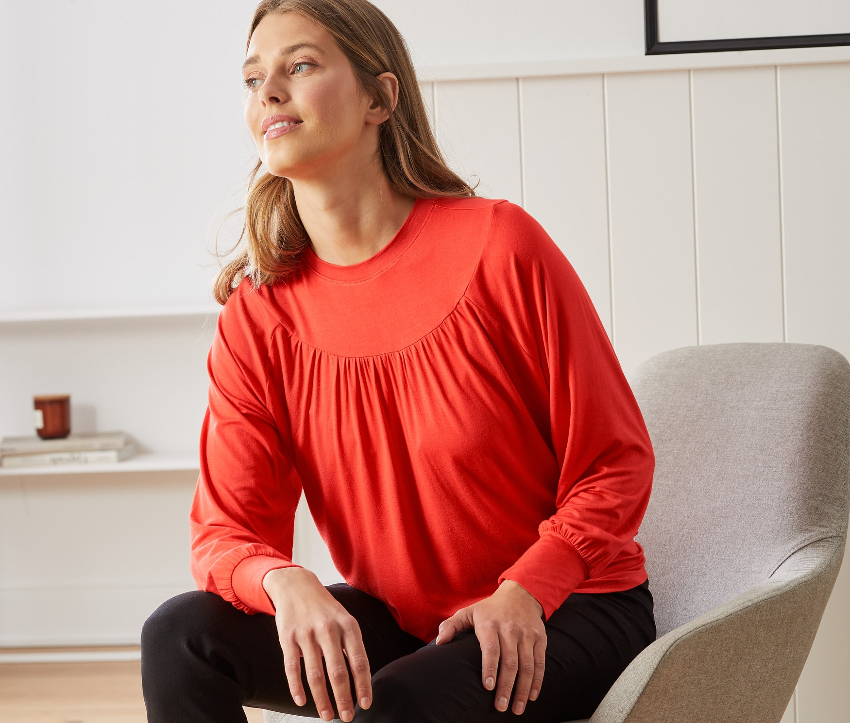 Bir kadın sandalyede oturuyor ve turuncu-kırmızı büzgülü bir bluz giyiyor.