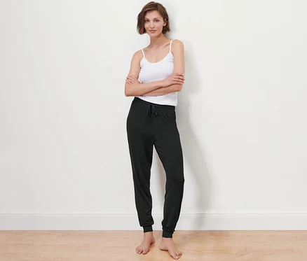 Beyaz bir duvarın önünde poz veren, atlet ve pijama altı giyen bir kadın.
