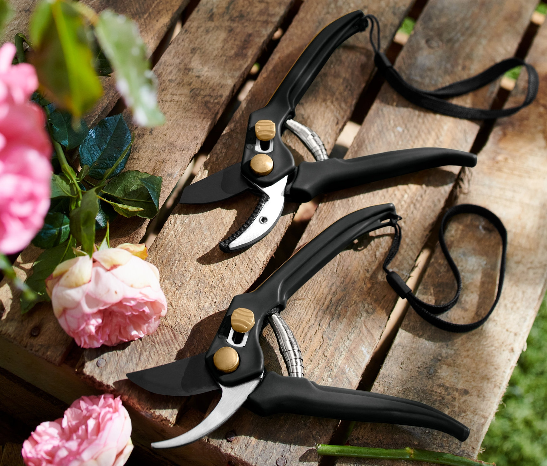 İki adet siyah bahçe makası, pembe güllerin yanında ahşap bir palet üzerinde duruyor. Bahçe Makası Seti.