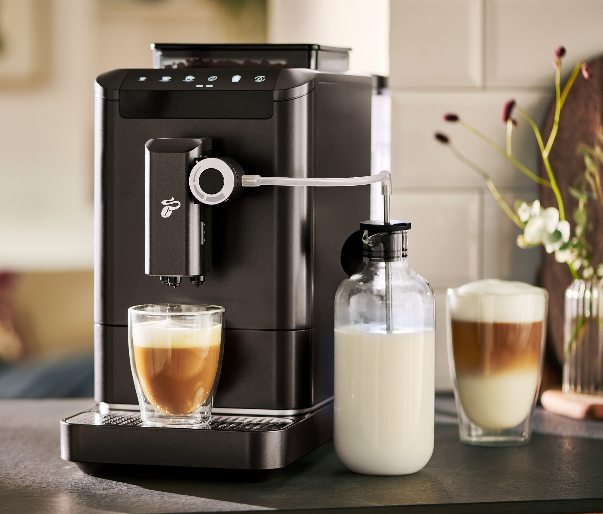 Bir »Esperto 2 Milk« Granite Black tam otomatik kahve makinesi bir bardağa kahve hazırlıyor. Yanında bir sütlük bulunmaktadır.