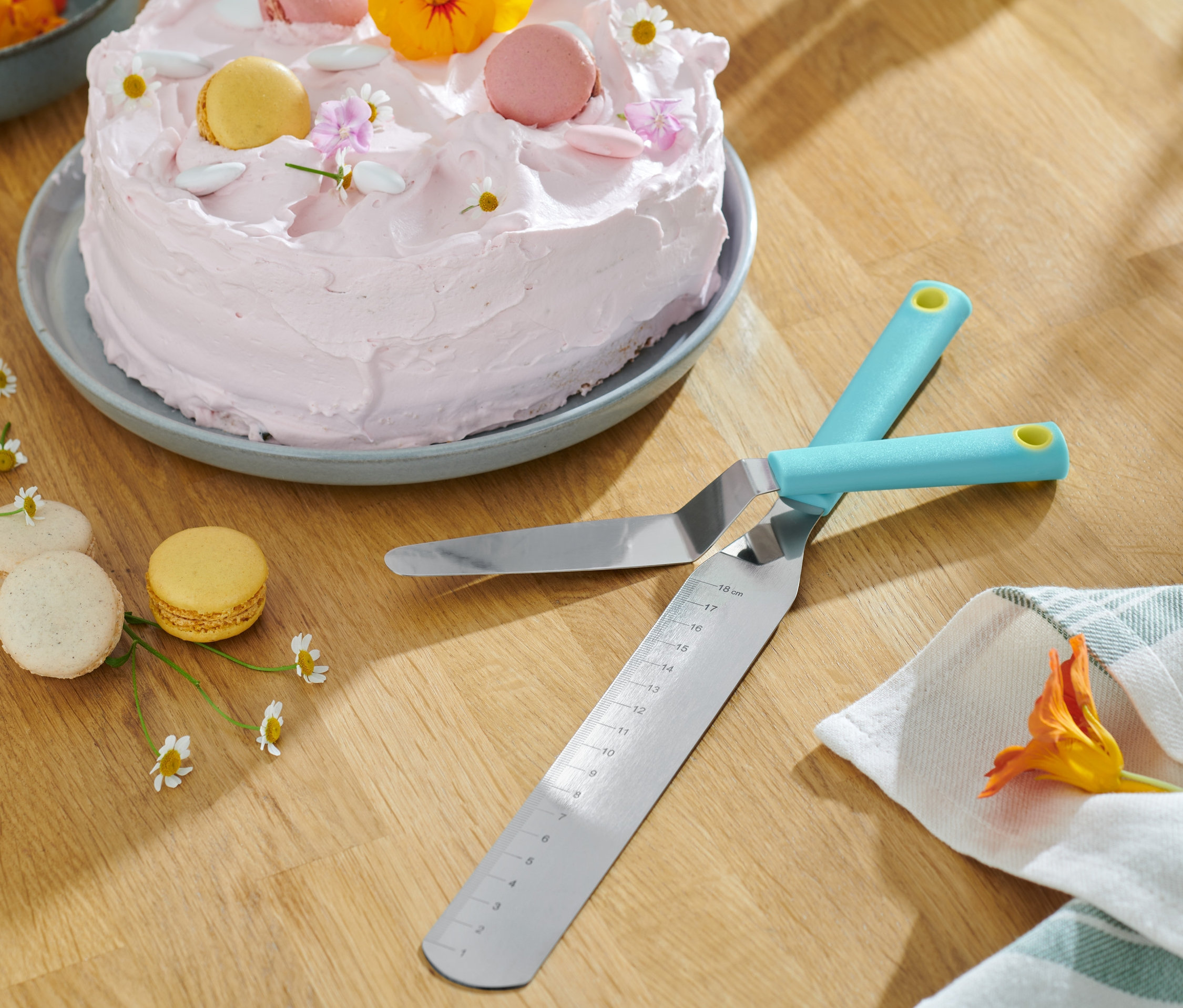 Ahşap bir masa üzerinde pembe krema ve makaronlarla süslenmiş bir pastanın yanında çapraz duran iki adet 2 Adet Pasta Spatulası.